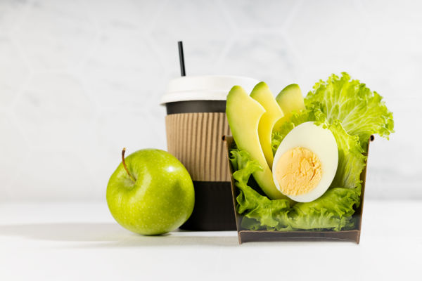 A cup filled with avocado, egg, and lettuce, with apple and coffee cup on the side.