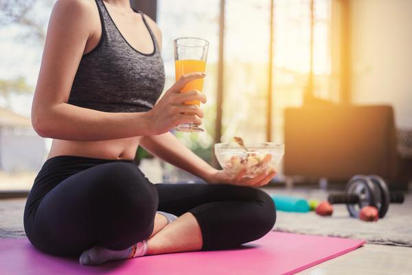 A woman sitting in workout clothes holding a glass of juice and a bowl of fruits