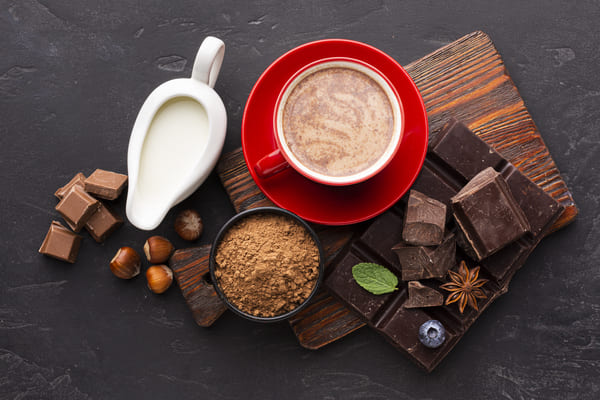 A cup of hot chocolate with milk, cocoa powder, chocolates, and hazelnuts