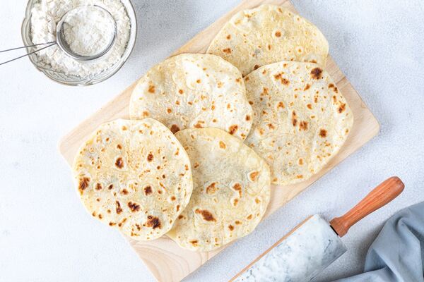 Homemade tortilla wraps on a wooden board with a bowl of flour on the side