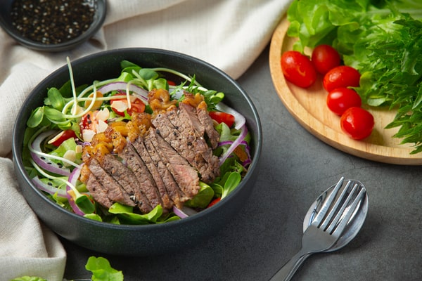 Slices of steak with colorful veggies served on a bed of greens and placed in a black bowl
