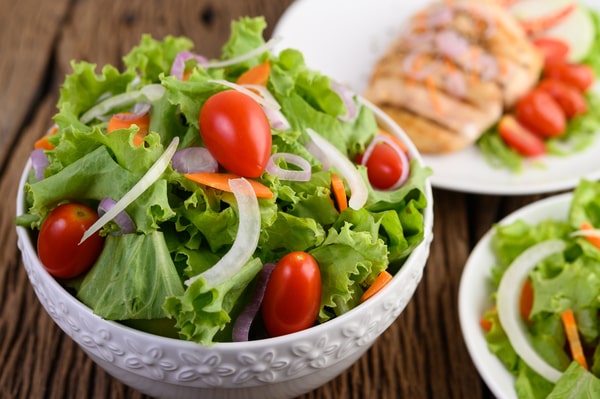 A bowl of fresh salad with a plate of grilled chicken on the background