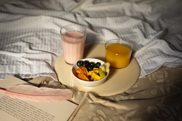 19 Homemade Drinks That Burn Fat While Sleeping: Find Out! 2 A cozy breakfast in bed featuring a plate with assorted fresh fruits, a glass of pink smoothie, a glass of orange juice, and an open book with a pink feather bookmark