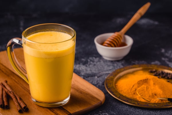 19 Homemade Drinks That Burn Fat While Sleeping: Find Out! 11 A glass mug of golden turmeric milk placed on a wooden board, accompanied by a plate of turmeric powder, cinnamon sticks, and a bowl of honey with a honey dipper, set against a dark background
