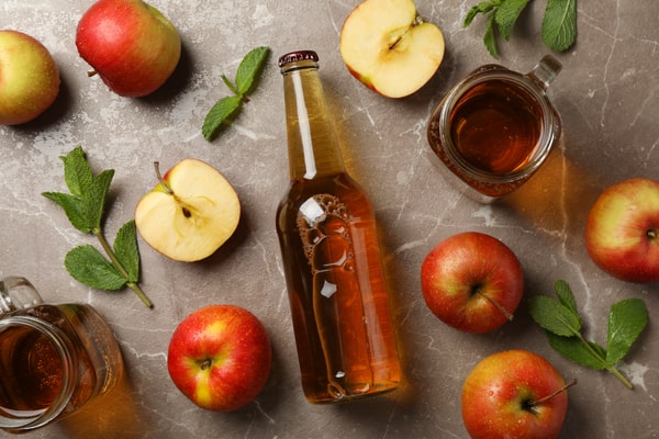 19 Homemade Drinks That Burn Fat While Sleeping: Find Out! 5 A bottle of apple cider surrounded by fresh apples, apple halves, mint leaves, and mugs of cider, arranged on a marble countertop