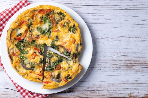Mushroom Frittata Recipe: Quick and Easy Meal You’ll Love 6 An Italian frittata made with spinach, tomatoes, onions, and peppers, served on a white plate with one slice removed, placed on a rustic white wooden table with a red checkered napkin