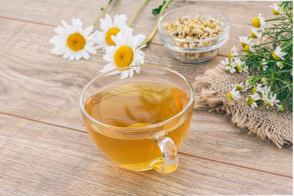 19 Homemade Drinks That Burn Fat While Sleeping: Find Out! 6 A glass cup of chamomile tea placed on a wooden table, surrounded by fresh chamomile flowers and a small glass bowl of dried chamomile blossoms