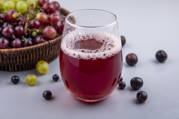 19 Homemade Drinks That Burn Fat While Sleeping: Find Out! 8 A glass of fresh grape juice with a basket of assorted grapes in the background, placed on a gray surface