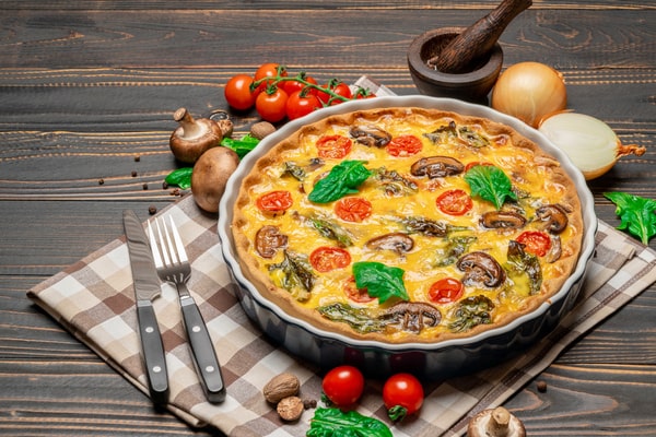 Mushroom Frittata Recipe: Quick and Easy Meal You’ll Love 5 A freshly baked homemade frittata mushrooms with cherry tomatoes, and spinach, presented in a ceramic baking dish on a checkered napkin, surrounded by fresh ingredients on a rustic wooden table
