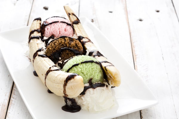 A classic banana split dessert with scoops of strawberry, chocolate, and mint ice cream, drizzled with chocolate syrup, and served on a white rectangular plate