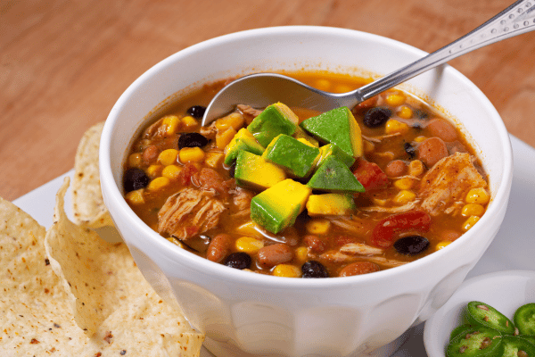 Keto Taco Soup Recipe: The Perfect Low Carb Comfort Food 2 Delicious bowl of chicken taco soup garnished with avocado with sides of chili peppers, sour cream, and tortillas.