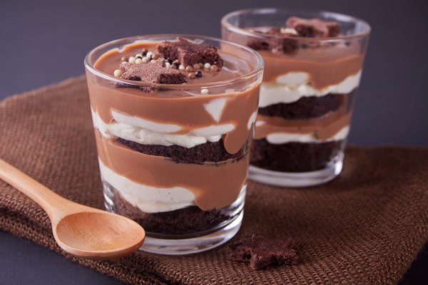 Two glasses of layered chocolate and cream dessert topped with chocolate crumbles and sprinkles, placed on a brown cloth next to a wooden spoon