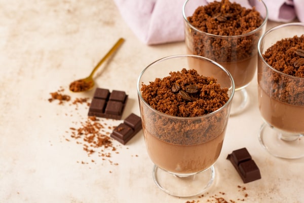 Three glasses of chocolate mousse topped with crumbled brownie, placed on a light stone surface with pieces of chocolate and a golden spoon nearby