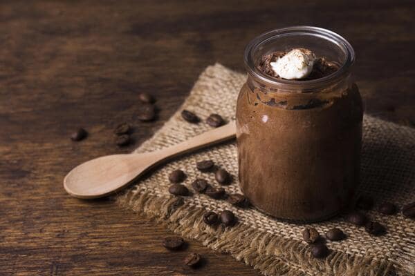 A jar of rich chocolate dessert topped with a dollop of cream, placed on a burlap cloth with coffee beans scattered around and a wooden spoon beside it on a rustic wooden surface