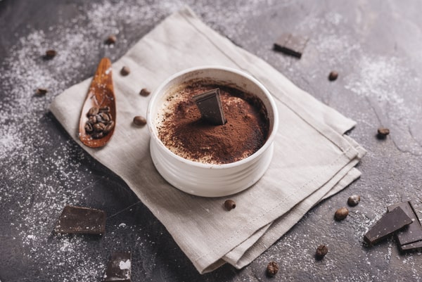
A white ceramic bowl filled with chocolate mousse, garnished with a square of dark chocolate and cocoa powder, placed on a folded cloth napkin