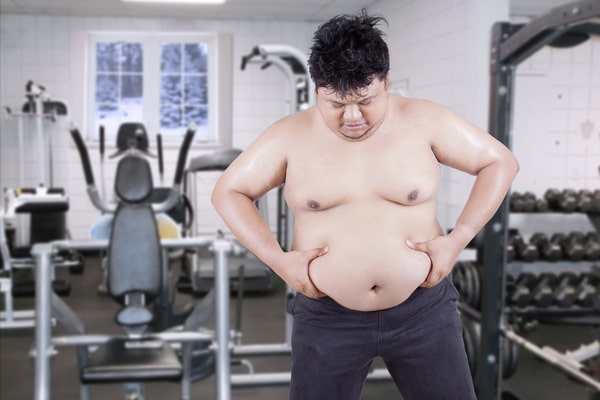 Signs of Gaining Muscle and Losing Fat: Quick and Easy Tips 9 An overweight man standing shirtless in a gym, grabbing his belly fat while looking down