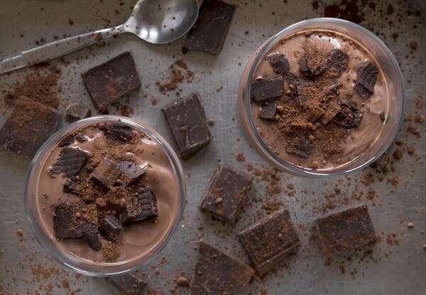 Top view of two glasses filled with creamy chocolate mousse topped with chocolate chunks and cocoa powder, surrounded by pieces of dark chocolate and a spoon