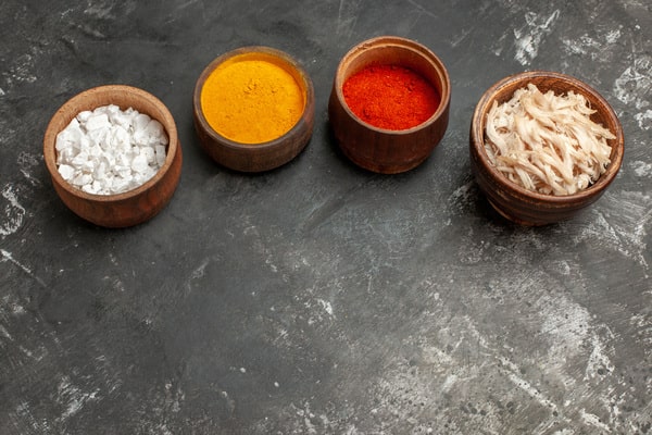 Four wooden bowls filled with different ingredients including salt, turmeric, red chili powder, and shredded ginger are arranged in a row on a dark grey background