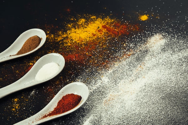 Three white ceramic spoons filled with different spices, including cinnamon, white pepper, and paprika, are placed diagonally on a black surface sprinkled with various colorful spices