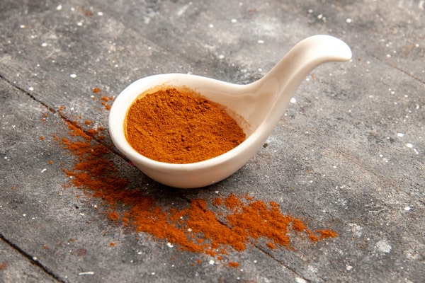 A white ceramic spoon filled with ground red pepper sits on a grey rustic surface with some pepper spilled around it