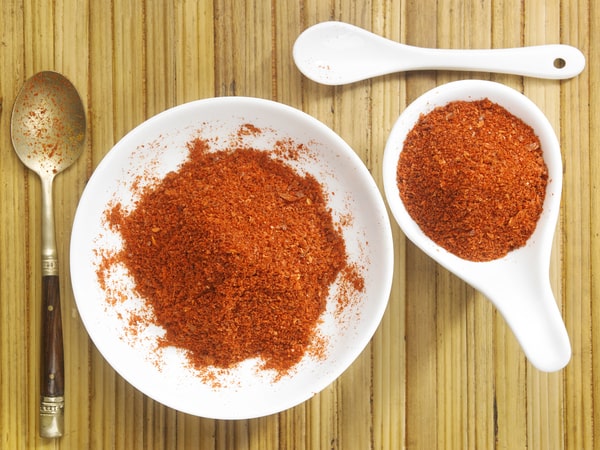 A top view of a white bowl and a white spoon filled with red chili powder, placed on a wooden table alongside a metal spoon and another ceramic spoon