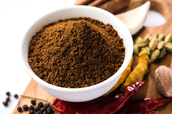 A bowl filled with garam masala powder is surrounded by various whole spices, including cardamom pods, dried red chilies, turmeric roots, and black peppercorns, on a wooden surface