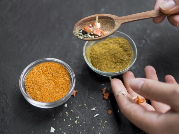 A pair of hands is holding a wooden spoon and some dried herbs, next to two glass bowls filled with different spices on a dark surface