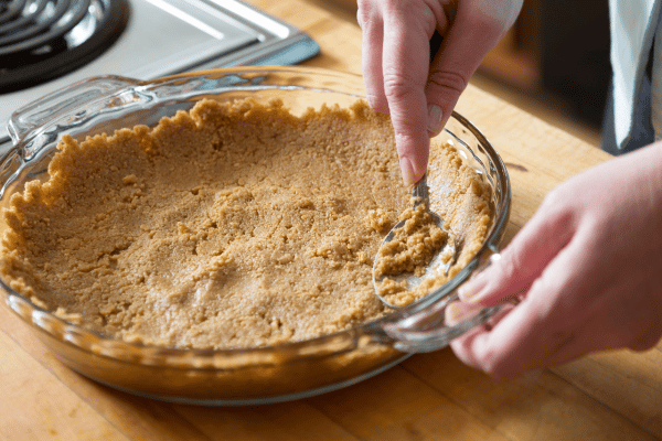A person gently pressing crushed graham crackers into a clear casserole dish with a spoon, creating an even layer for the base.