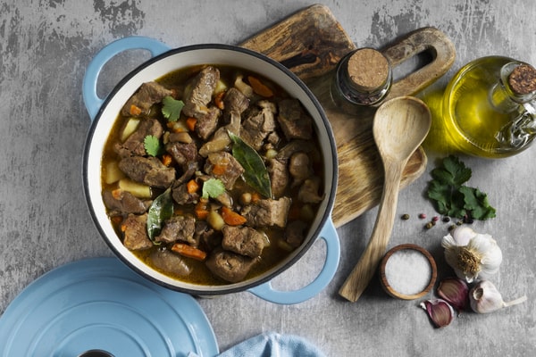 Keto Pot Roast: A Hearty and Healthy Meal Option! 11 blue Dutch oven filled with aromatic beef stew, carrots, potatoes, and bay leaves, surrounded by wooden utensils, olive oil, garlic, salt, and herbs on a rustic wooden board and textured gray surface