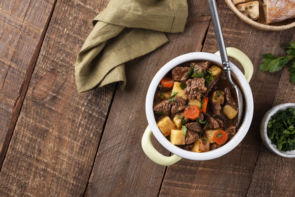 Keto Pot Roast: A Hearty and Healthy Meal Option! 3 A cozy bowl of beef pot with chunks of meat, potatoes, and carrots garnished with herbs, placed on a rustic wooden table alongside a folded green napkin, bread, and fresh parsley