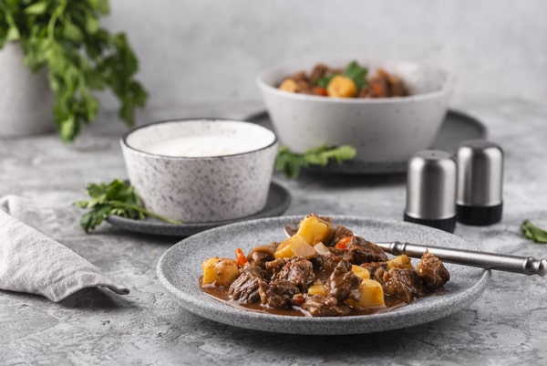 Keto Pot Roast: A Hearty and Healthy Meal Option! 5 A gray plate filled with savory beef stew, potatoes, and carrots, accompanied by bowls of yogurt and stew in the background, garnished with fresh herbs