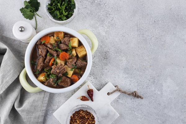 Keto Pot Roast: A Hearty and Healthy Meal Option! 7 A pot roast beef stew with chunks of meat, carrots, and potatoes garnished with fresh herbs, set on a light gray surface with a marble board holding chili flakes and dried chili, alongside a napkin and small bowls of parsley