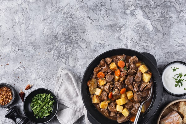 Keto Pot Roast: A Hearty and Healthy Meal Option! 8 A cast iron pot filled with rich beef stew, potatoes, and carrots, surrounded by bowls of fresh parsley, chili flakes, and yogurt, along with a napkin and crusty bread on a textured gray surface