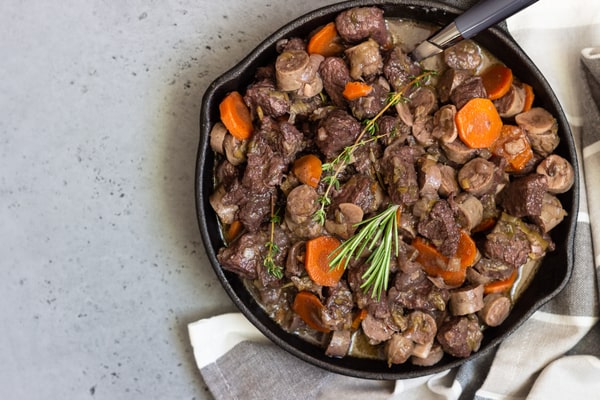 Keto Pot Roast: A Hearty and Healthy Meal Option! 9 A cast iron skillet filled with beef roast featuring tender chunks of beef, sliced sausages, carrots, and mushrooms, garnished with fresh rosemary and thyme, set on a gray surface with a striped cloth napkin