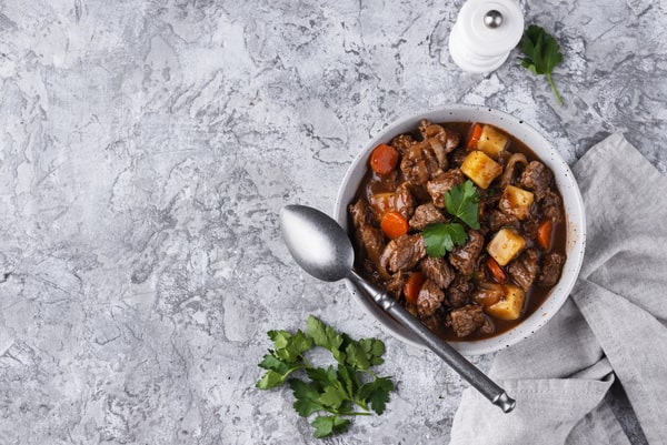 Keto Pot Roast: A Hearty and Healthy Meal Option! 10 A bowl of flavorful beef stew with tender meat, potatoes, and carrots, garnished with fresh parsley, placed on a textured gray surface with a napkin, a spoon, and a white pepper grinder nearby
