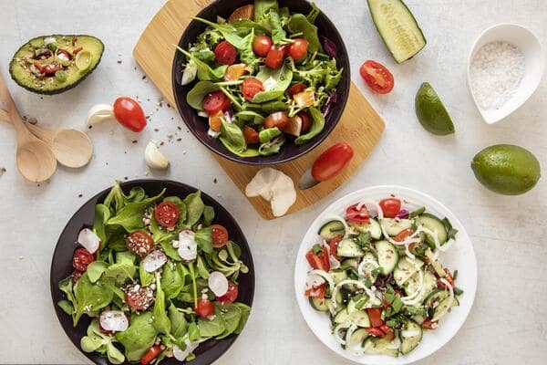 10 Low Carb Salads Perfect for Summer Picnics 3 Three bowls of low-carb salads made with lettuce, cucumber, tomatoes, and radishes, placed on a table with fresh veggies, lime, and garlic