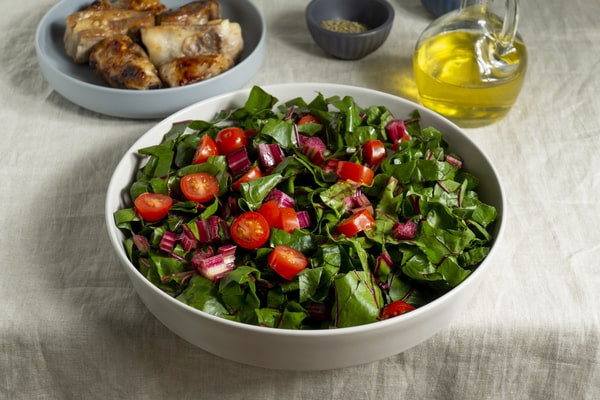 10 Low Carb Salads Perfect for Summer Picnics 4 A low-carb salad made with chopped leafy greens, cherry tomatoes, and purple stems, served in a white bowl with grilled meat and olive oil nearby