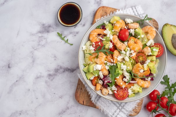 10 Low Carb Salads Perfect for Summer Picnics 8 A low-carb salad with shrimp, cherry tomatoes, avocado, lettuce, and crumbled cheese, served in a bowl on a marble table with dressing on the side