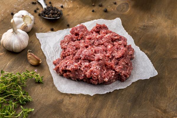 Calories in Ground Beef: Health Facts You Should Know 2 A mound of raw ground beef sits on parchment paper surrounded by fresh herbs, garlic, peppercorns, and a spoon on a wooden surface