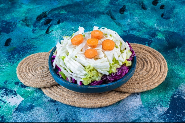 Easy Red White and Blue Salad Recipe for Your BBQ 11 A blue bowl of red white and blue salad with shredded cabbage, purple cabbage, lettuce, and carrot slices on a woven trivet