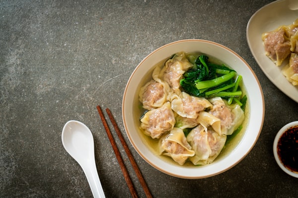 21 Keto Chinese Recipes: Enjoy Takeout Without the Carbs 13 A top-down view of a bowl of wonton soup with dumplings and green vegetables, with chopsticks and a spoon resting beside it.
