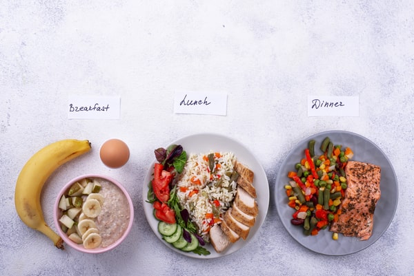 3 Day Keto Diet: Simple Steps to Jumpstart Ketosis 3 A flat lay of three balanced meals labeled breakfast, lunch, and dinner with fruit, oatmeal, chicken with rice and salad, and salmon with mixed vegetables