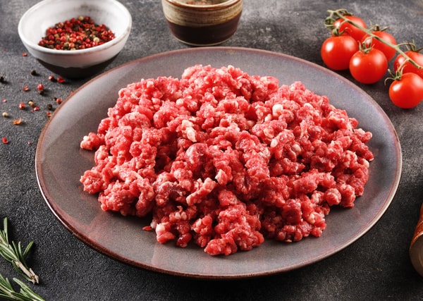 Calories in Ground Beef: Health Facts You Should Know 3 A plate of raw ground beef is displayed on a dark surface, surrounded by ingredients like cherry tomatoes, rosemary, and peppercorns