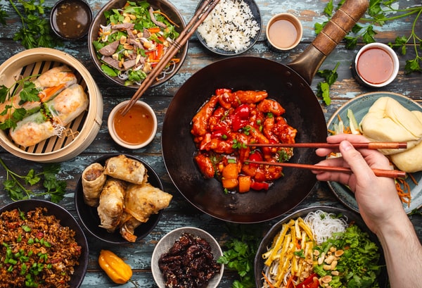 21 Keto Chinese Recipes: Enjoy Takeout Without the Carbs 3 A person uses chopsticks to take a piece of sweet and sour chicken from a large wok in the center of a table laden with various Chinese dishes.
