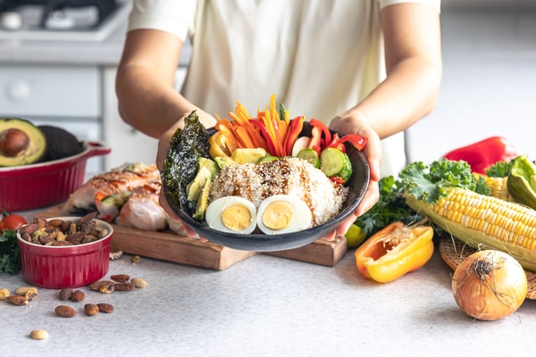How Do You Start the Keto Diet? 5 Steps for Beginners 4 A person holding a bowl of mixed vegetables, boiled eggs, avocado, and rice surrounded by fresh produce and nuts on a kitchen counter