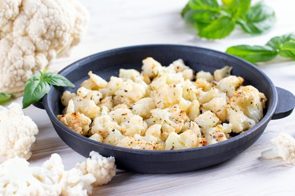 Cauliflower Bake: Deliciously Cheesy and Healthy Recipe 5 Roasted cauliflower pieces seasoned with black pepper in a cast-iron pan, surrounded by fresh basil leaves