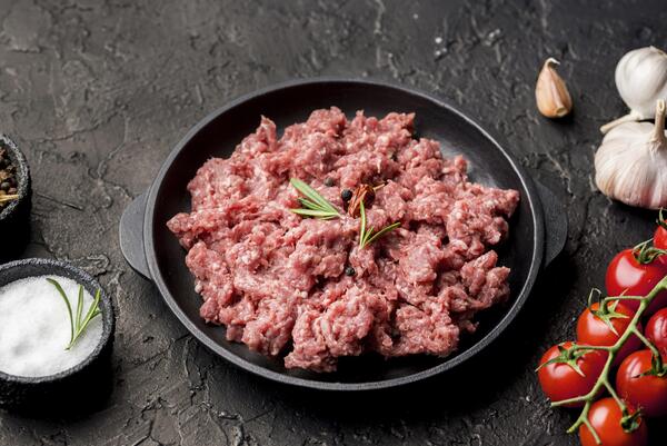 Calories in Ground Beef: Health Facts You Should Know 6 Raw ground beef garnished with rosemary is served in a black dish, surrounded by cherry tomatoes, garlic, salt, and pepper