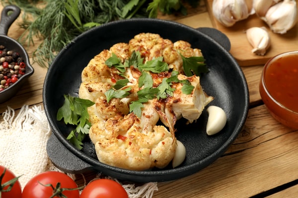 Cauliflower Bake: Deliciously Cheesy and Healthy Recipe 6 A roasted cauliflower steak garnished with fresh parsley, surrounded by garlic cloves, tomatoes, and spices on a wooden table