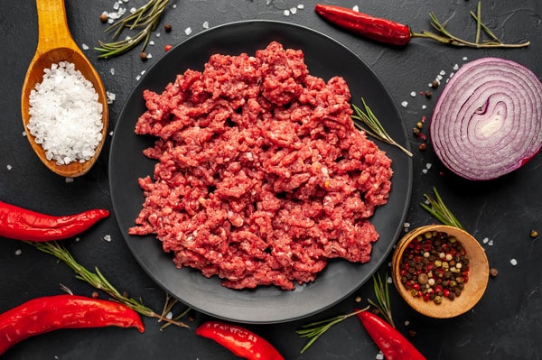 Calories in Ground Beef: Health Facts You Should Know 7 A black plate filled with raw ground beef is surrounded by coarse salt, red chili peppers, rosemary, peppercorns, and a halved red onion