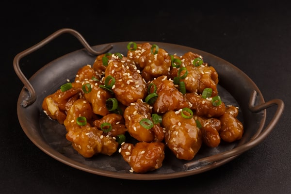 21 Keto Chinese Recipes: Enjoy Takeout Without the Carbs 7 A dark metal plate with handles holds a mound of General Tso's chicken, garnished with sesame seeds and chopped scallions.